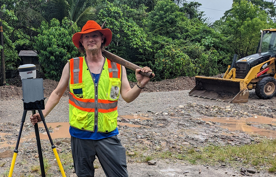 Barr employee Gareth Becker wearing a bright safety vest and hat posing with surveying equipment while volunteering in Peru