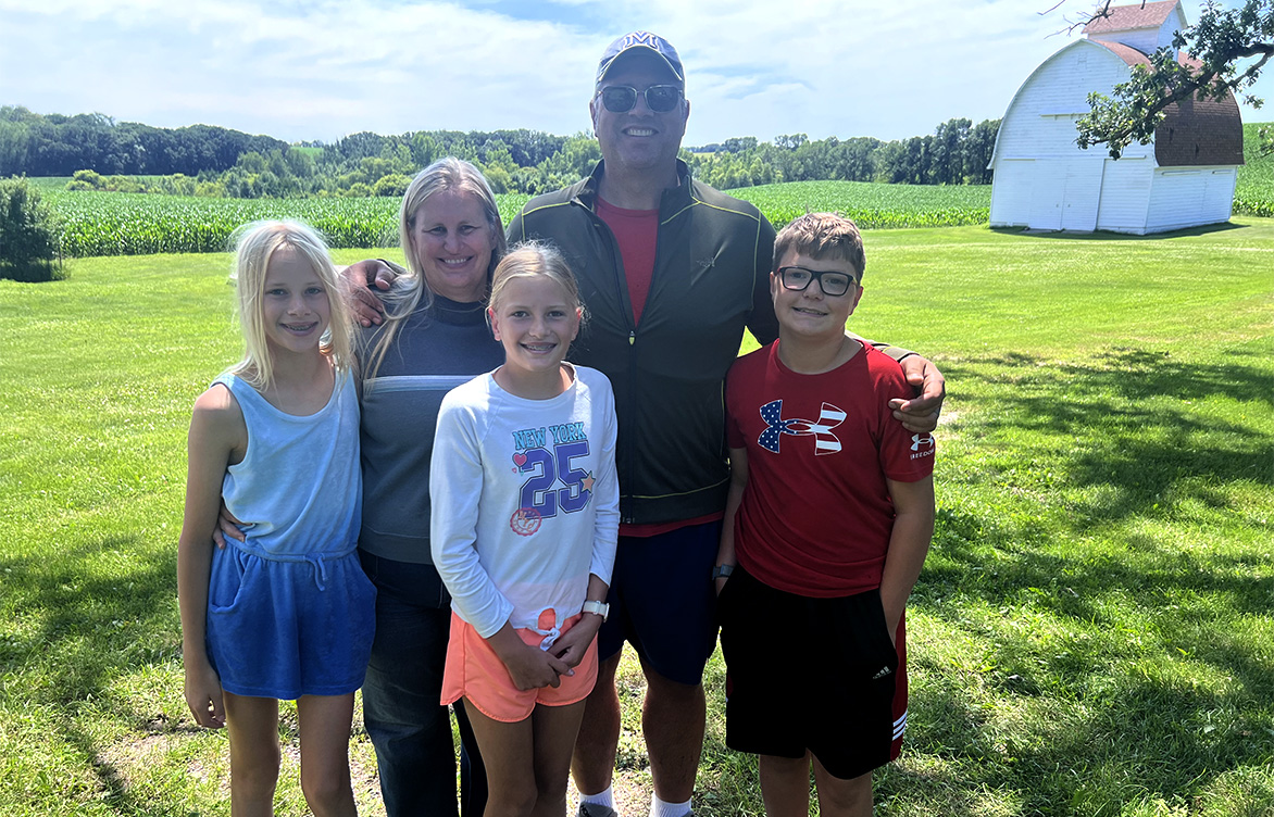 Jason, his wife, two daughters, and son pose for a photo in front of a cornfield.