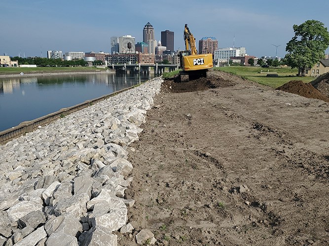 An excavator working on a levee along a river with a city skyline and bridge in the background.