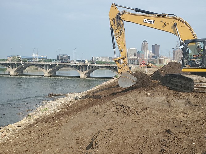 An excavator working on a levee along a river with a city skyline and bridge in the background.