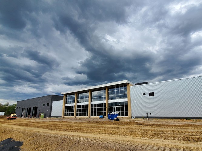 A large industrial building with tall glass windows sits on a dirt construction site under dark, dramatic storm clouds.