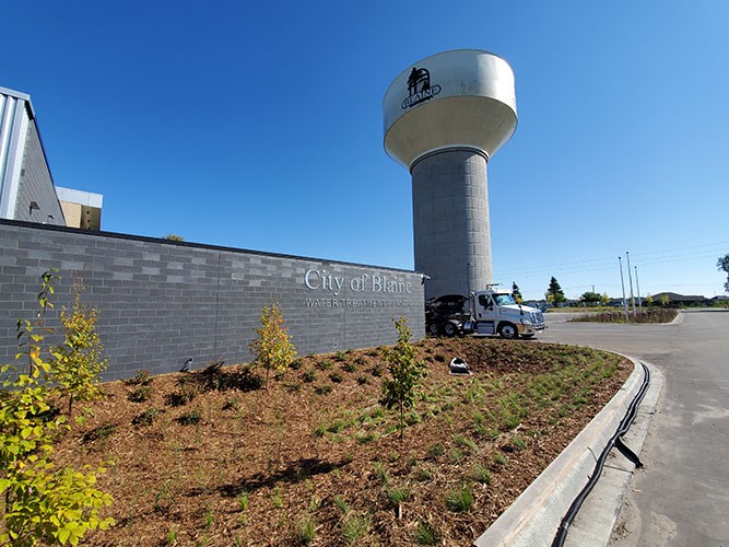 A newly planted landscape with young trees and mulch sits beside a gray brick building labeled “City of Blaine Water Treatment Plant” with a large water tower rising behind it.