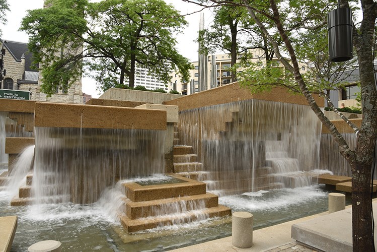 A tiered concrete fountain with multiple cascading waterfalls flows into shallow pools, creating stepped terraces of water. Large trees surround the fountain, and modern buildings rise in the background.
