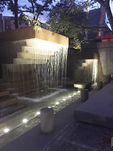 A softly lit tiered concrete fountain spills water into a narrow channel at dusk, with ground lights illuminating the flowing water and surrounding structures.