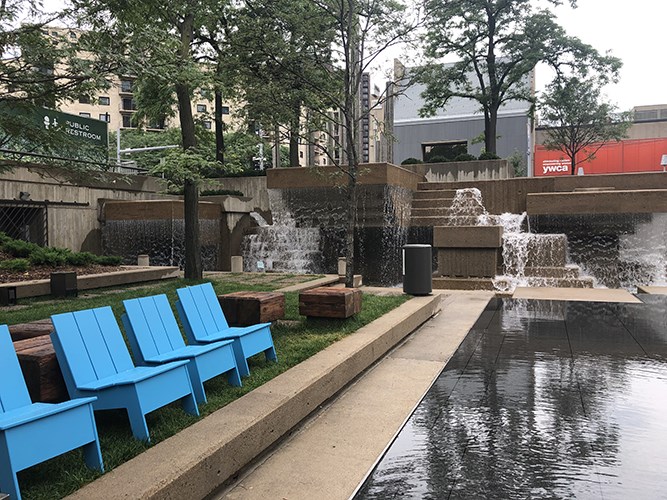 Bright blue chairs sit beside a reflective plaza near a tiered concrete fountain with cascading water, surrounded by trees and urban buildings.