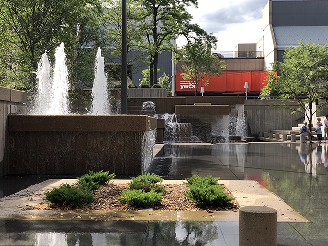 A terraced concrete fountain with multiple waterfalls sits beside a shallow reflective plaza, framed by trees and modern buildings with a red YWCA sign in the background.