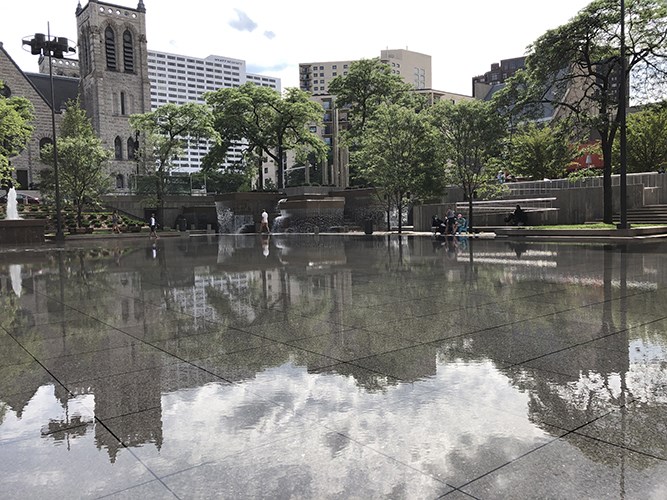 A shallow reflective plaza mirrors surrounding trees, buildings, and a tiered concrete fountain in an urban park setting.