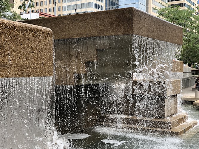A tiered concrete fountain spills sheets of water over its edges into a shallow pool, with modern buildings rising behind the structure.