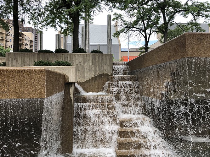 A stepped concrete fountain cascades down multiple tiers between high walls, framed by trees and urban buildings in the background.