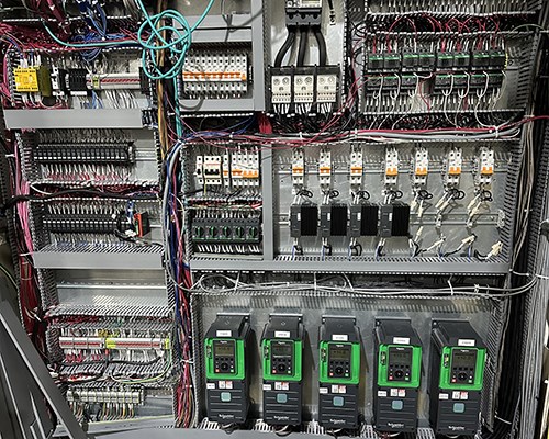Interior of an electrical control panel with circuit breakers, relays, terminal blocks, and wiring, including five green and black variable frequency drives at the bottom.