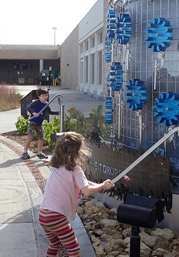 A child in the foreground uses a long handle to spin blue gear-like wheels on a cylindrical outdoor art piece while another child does the same nearby along the landscaped walkway.