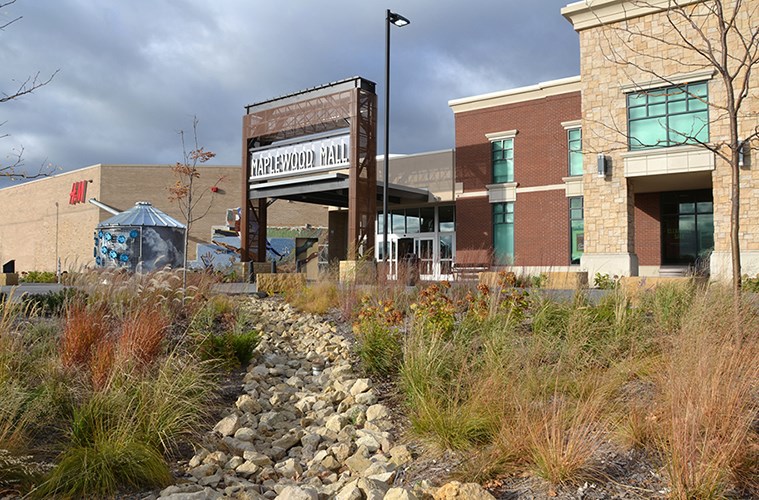 Exterior view of Maplewood Mall showing landscaped rocks and grasses in the foreground and modern brick-and-stone buildings under a cloudy sky.