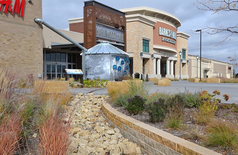 A landscaped shopping‑center entrance features a rocky drainage swale, ornamental grasses, and a small metal grain‑bin structure near storefronts including a bookstore.