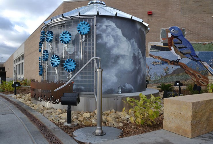 A cylindrical metal structure painted with storm‑cloud imagery and decorated with blue gear‑like shapes stands beside a bird mural and landscaped rocks.