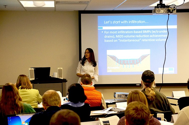 A person at the front of a classroom gives a presentation while attendees seated at tables watch a projected slide about infiltration practices.