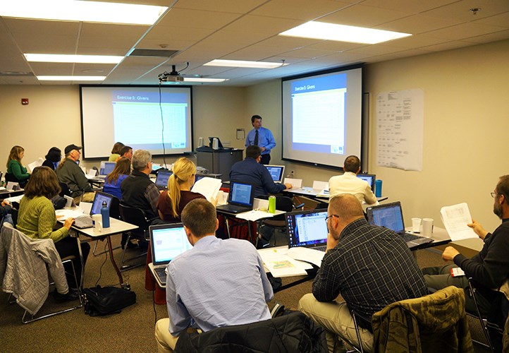 A classroom of people seated at tables with laptops and papers watches an instructor presenting slides at the front of the room.