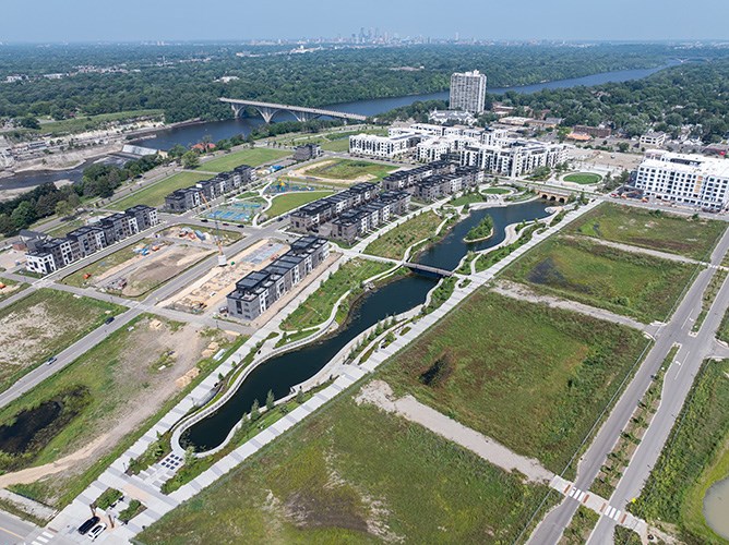 Aerial image of an urban, residential development site next to a large river with a long, narrow pond extending through the middle on a sunny day.