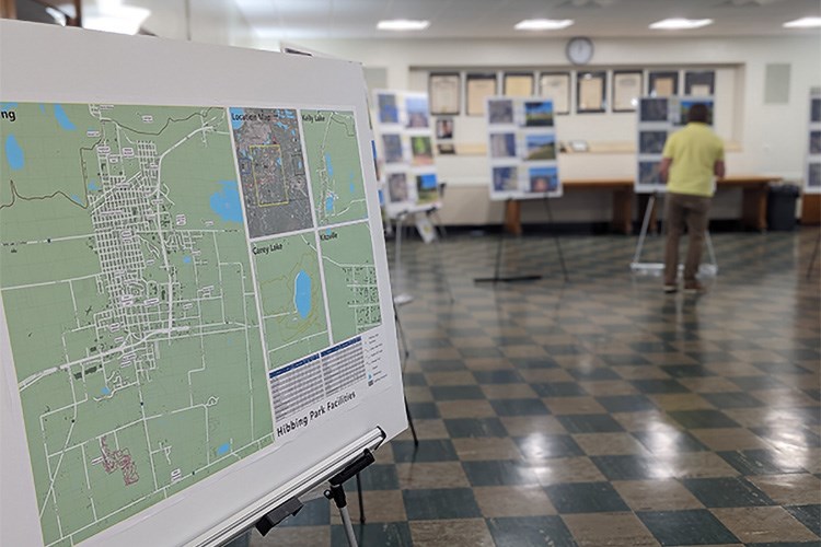 A poster-size map presented on an easel at a public meeting with a person examining other posters in the background.