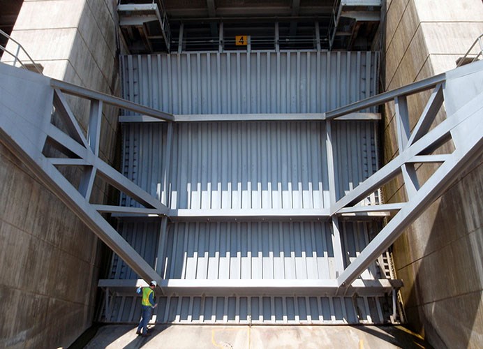Massive metal dam spillway gate with reinforced beams between concrete walls, marked with a yellow sign showing the number 4, and a person in safety gear standing at the bottom for scale.