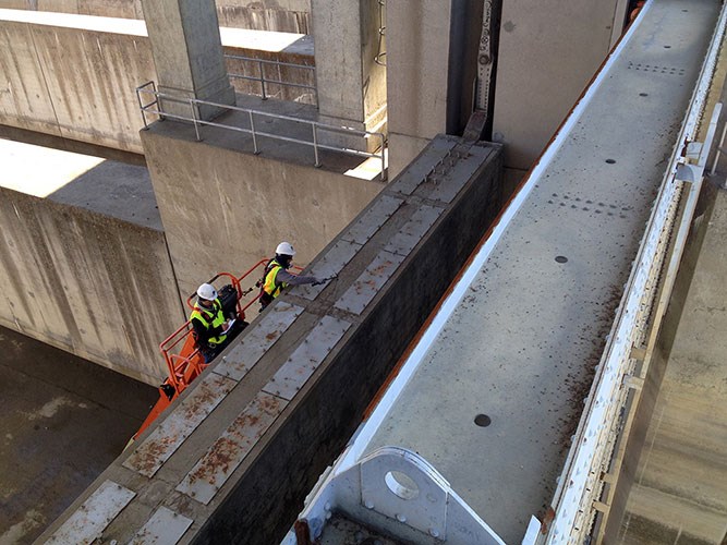 Two workers in helmets and high-visibility vests on an orange lift platform inspecting a large dam spillway gate.