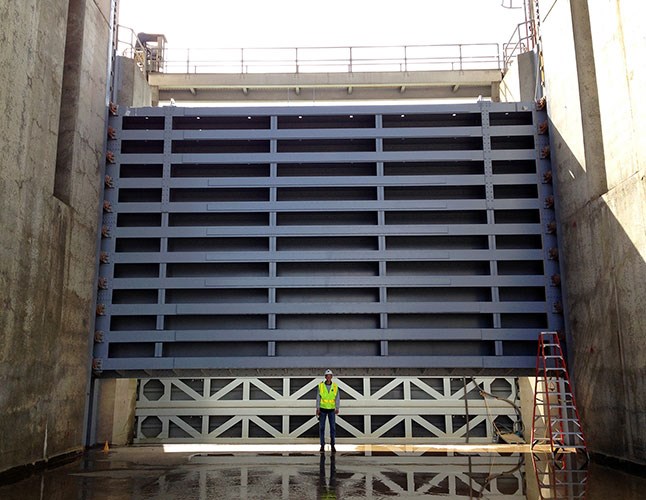 Person in safety gear standing in front of a massive metal spillway gate at a dam.