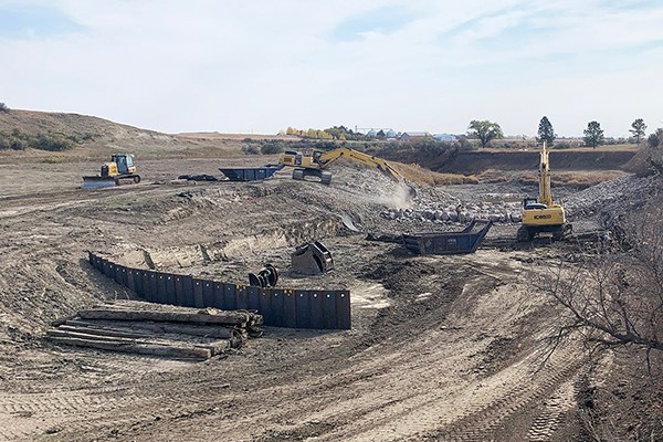 Excavators and construction equipment work in a wide, dusty excavation area with steel shoring panels and piles of rock scattered throughout the site.
