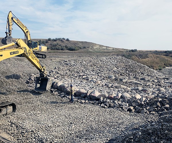Excavator positions large riprap boulders on a wide gravel slope while a worker stands nearby for scale.