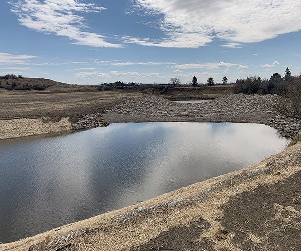 A small pond reflects the sky in a dry, grassy landscape bordered by wide slopes of riprap stones.