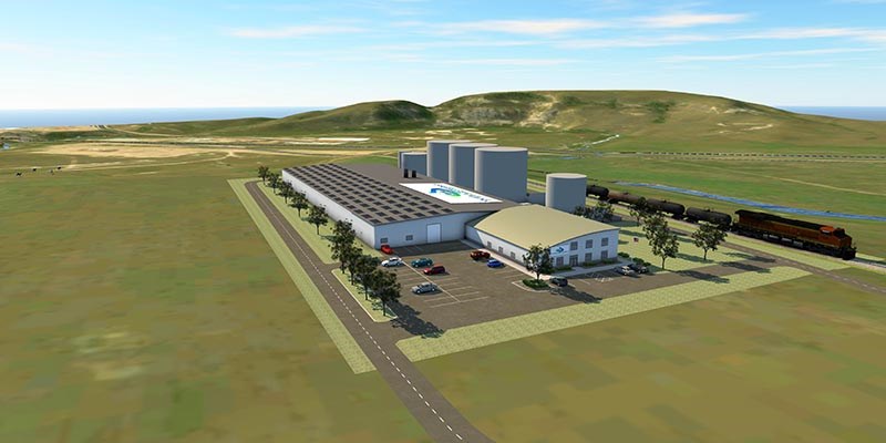 A large industrial facility with multiple long buildings, storage silos, a parking lot, and a nearby train sits in an open landscape of rolling green hills under a clear sky.