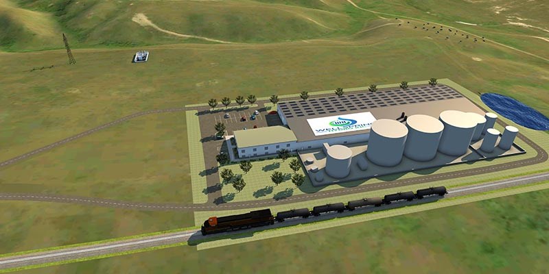 A large industrial facility with multiple long buildings, cylindrical storage tanks, a parking lot, and an adjacent rail line sits within a wide open landscape of grassy hills.