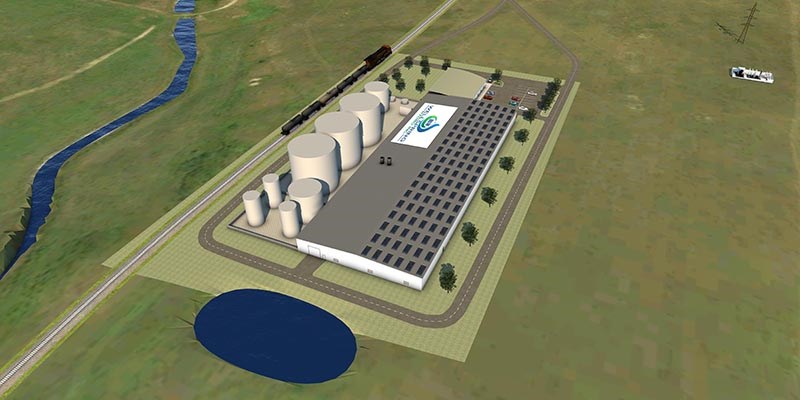 An industrial facility with large cylindrical storage tanks, long buildings, a parking lot, a nearby rail line, and a small pond sits within a wide open landscape of grassy hills.