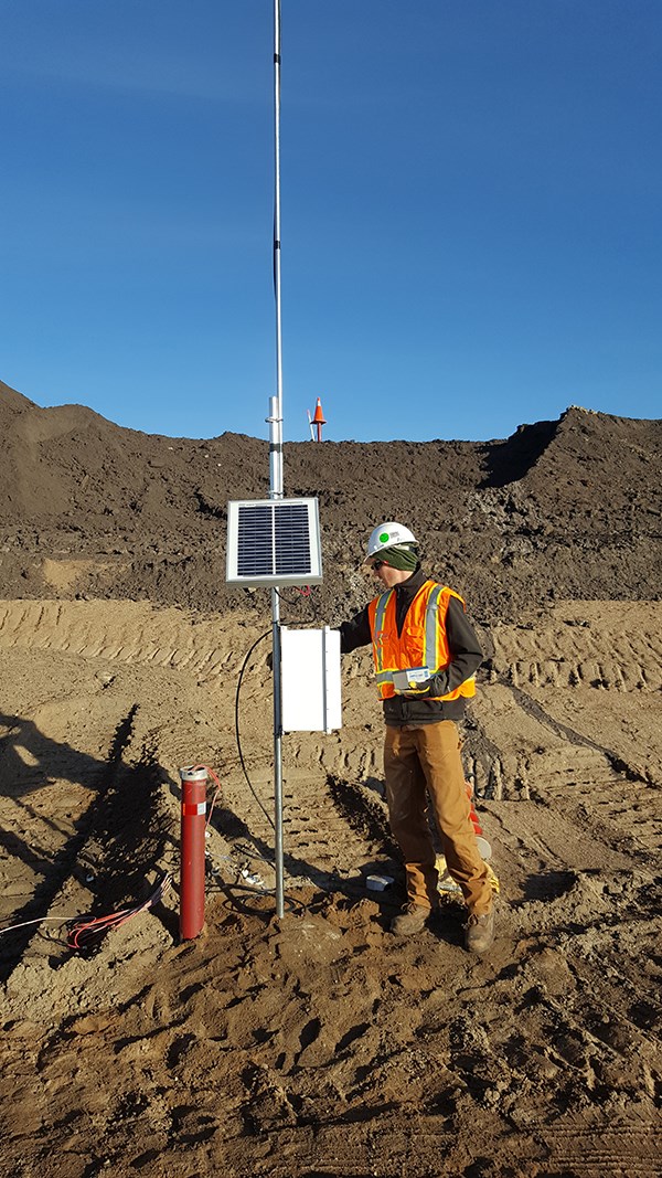 Person in safety gear adjusting a solar-powered monitoring device among dirt mounds and tire tracks under a clear blue sky.