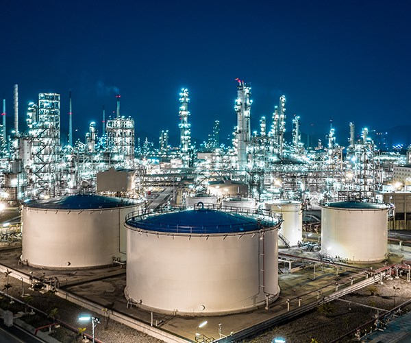 A brightly lit industrial refinery at night features large cylindrical storage tanks in the foreground and a dense array of illuminated towers and pipes in the background.