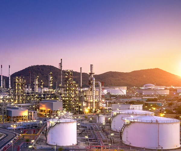 A large industrial refinery complex with cylindrical storage tanks sits in front of hills as the sun sets behind them, casting a warm glow over the facility.