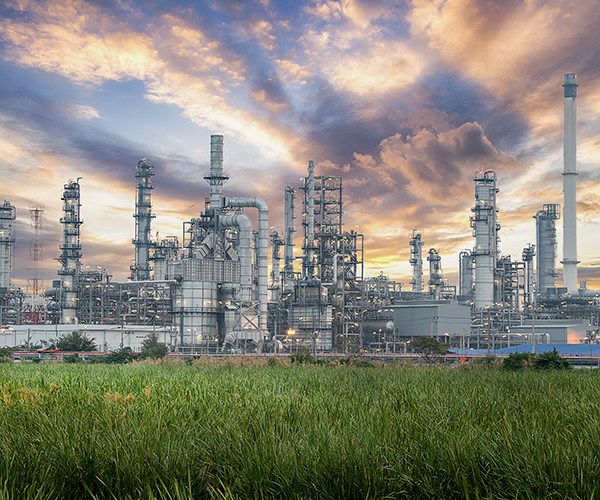 An industrial refinery complex with numerous tall metal towers and pipes rises behind a field of green grass under a dramatic, colorful sky.