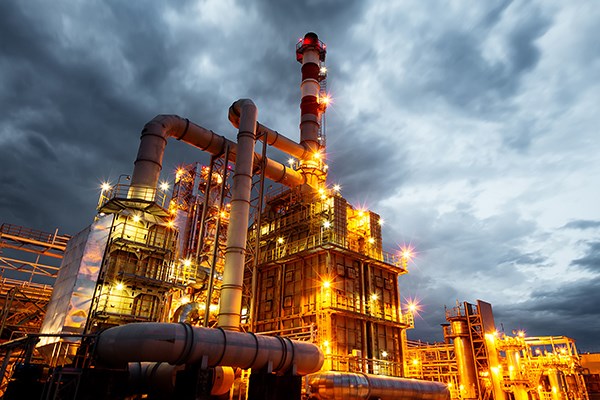 An illuminated industrial processing structure with large pipes and a tall stack glows orange against a dark, stormy sky.